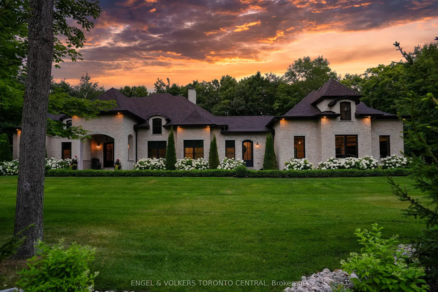 Beige brick house with a dark roof, green lawn, white flowers, and a sunset sky.
