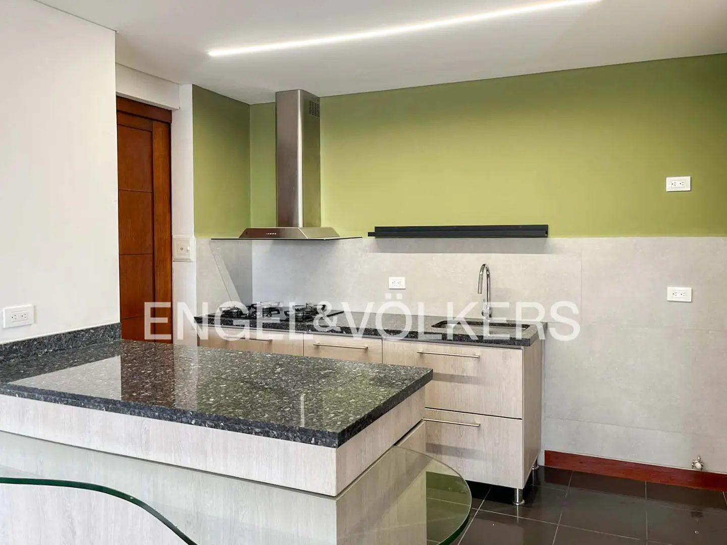 A modern kitchen with a black granite countertop, stainless steel hood, and light wood cabinets. The walls are green and gray.