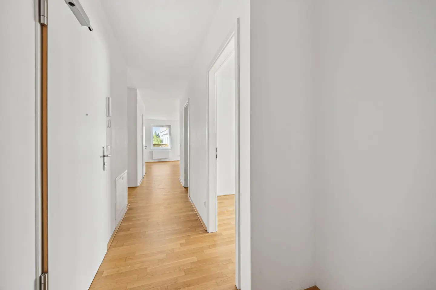 Bright, white hallway with light wood floors. A window is visible at the end of the hall.