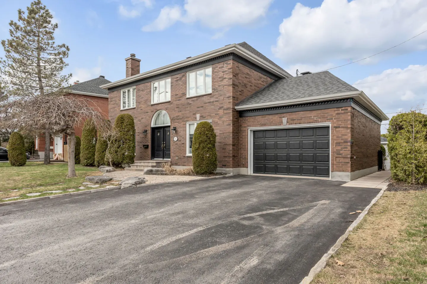 Two-story brick house with a black garage door and front door, set on a paved driveway with green bushes.