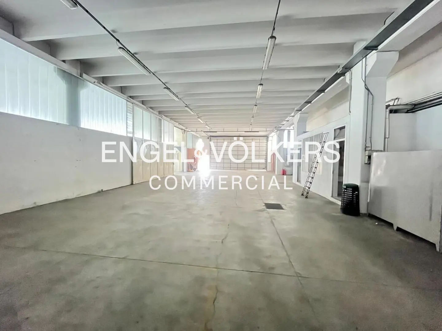 Large, empty commercial space with white walls, concrete floors, and fluorescent lighting. "Engel & Völkers Commercial" logo is superimposed.