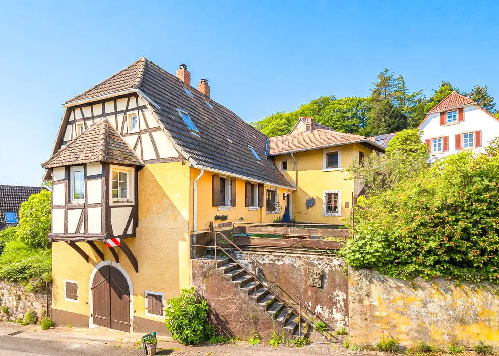 A yellow, half-timbered house with a brown door and a tiled roof under a blue sky. Steps lead to a balcony.