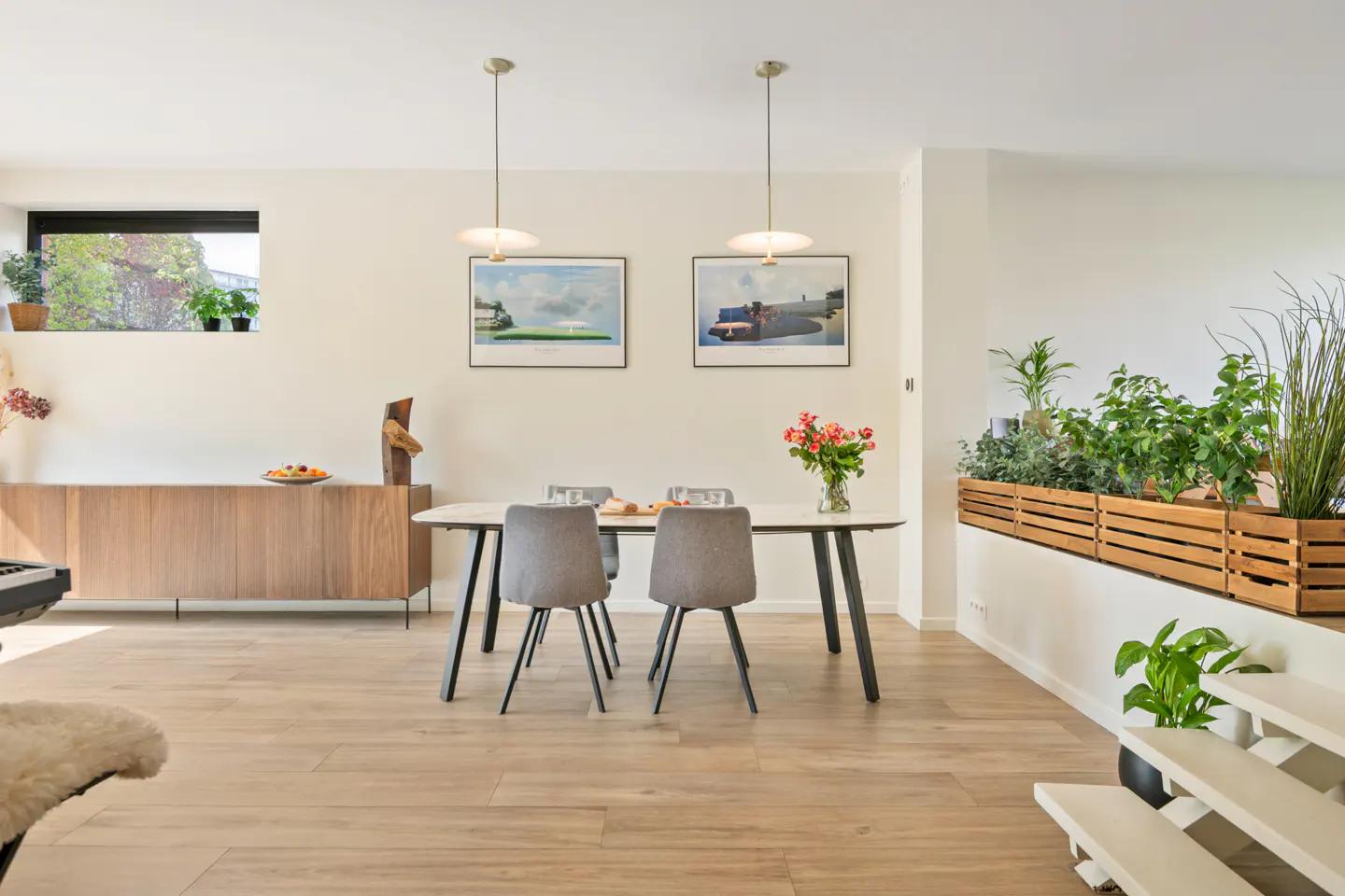 Bright dining room with wood floors, a white table with gray chairs, and a built-in planter with greenery. Two pendant lights hang above the table.