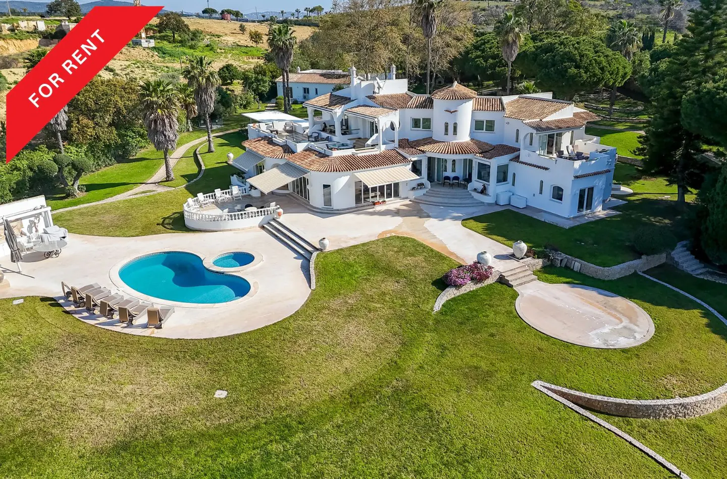 Aerial view of a large, white, Spanish-style villa with a pool and lush green lawn. Red "For Rent" banner in the upper left corner.