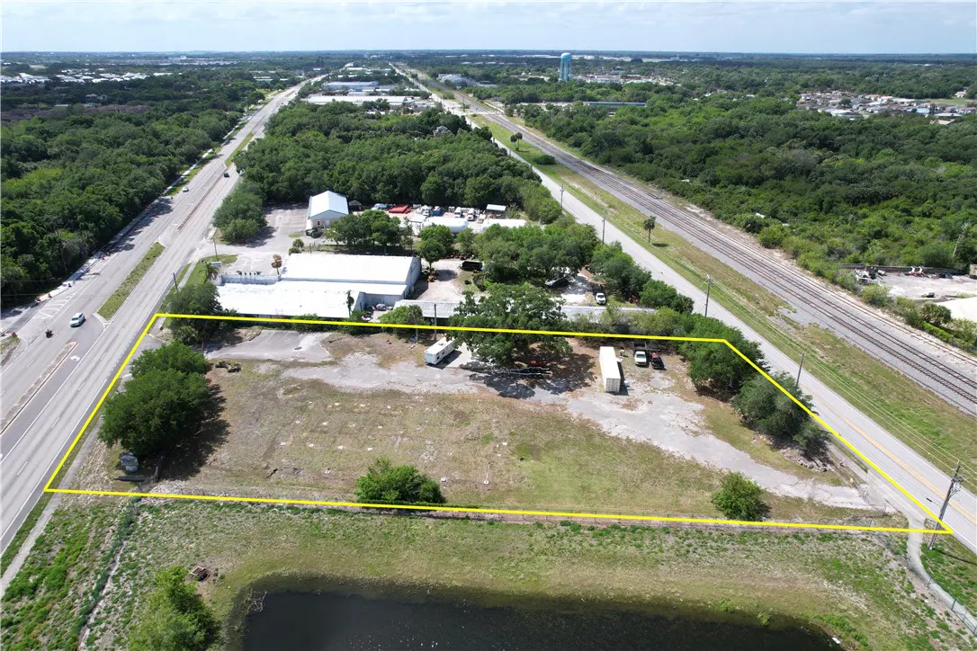 Aerial view of a vacant lot outlined in yellow, near a pond, highway, and railroad tracks. Buildings and trees surround the property.