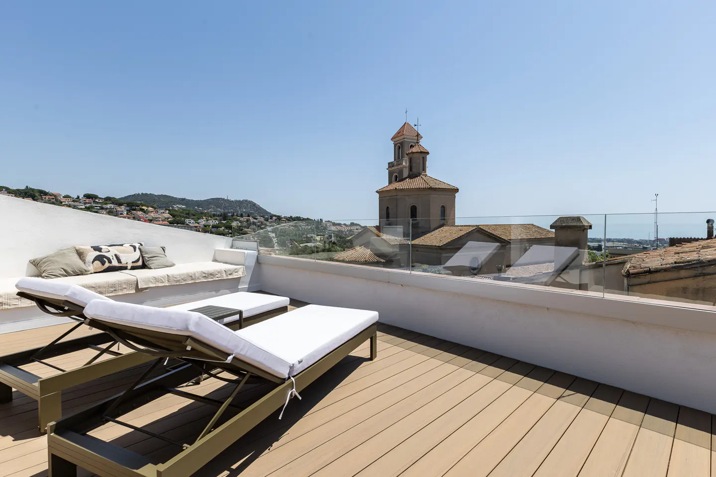 Rooftop terrace with two lounge chairs, a white bench with pillows, and a view of a church and town.