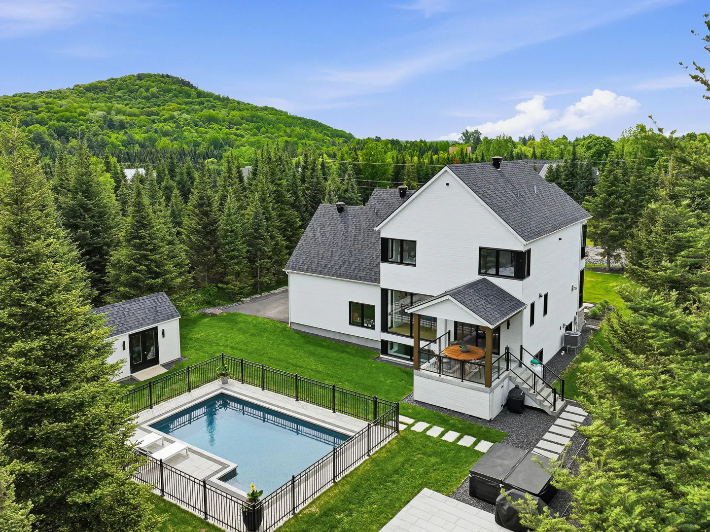 Aerial view of a modern white house with a pool, hot tub, and detached garage, surrounded by lush forest and mountains.