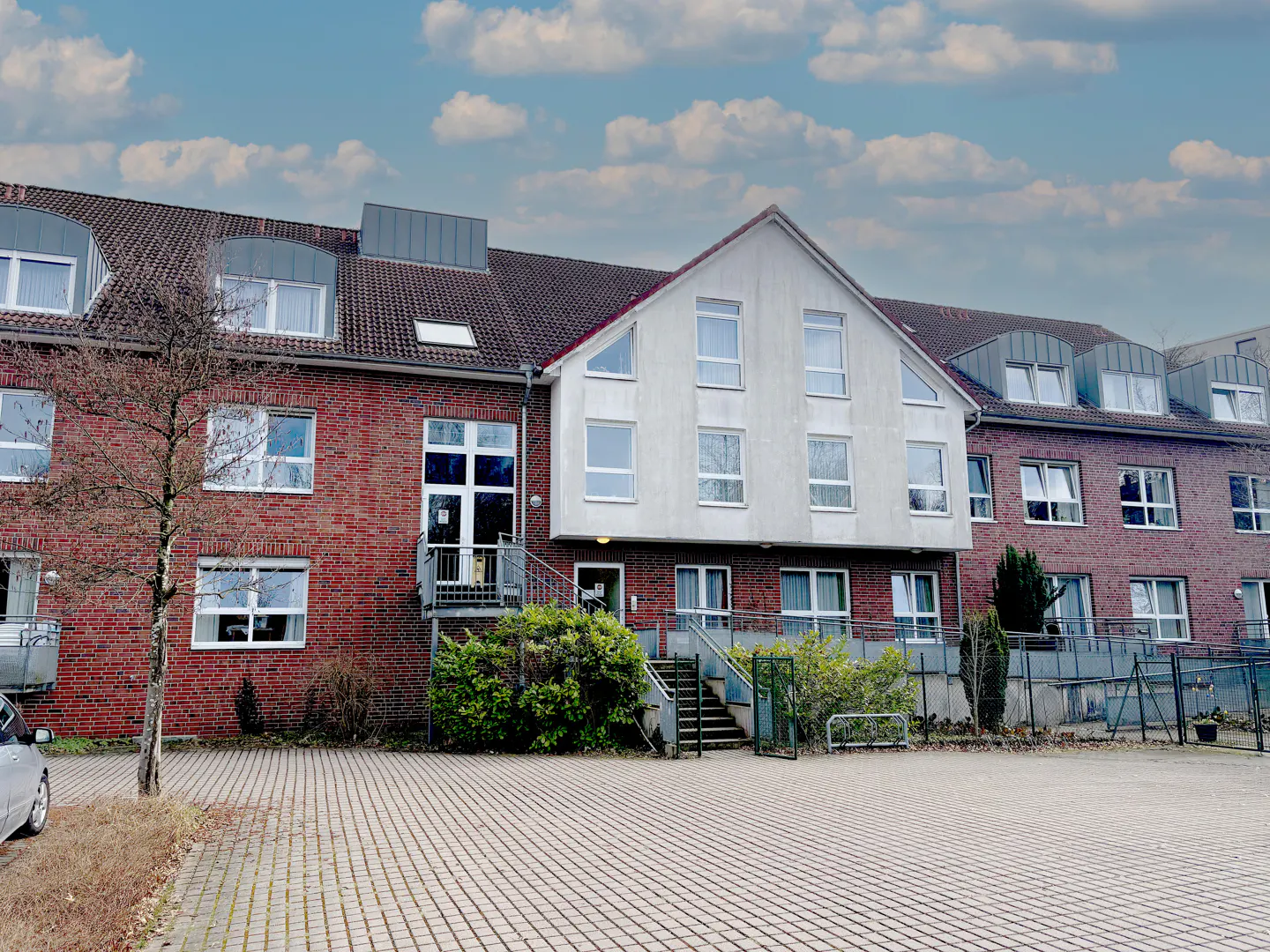 Exterior of a brick and white building with a tiled roof under a cloudy sky. A paved parking area is in front.