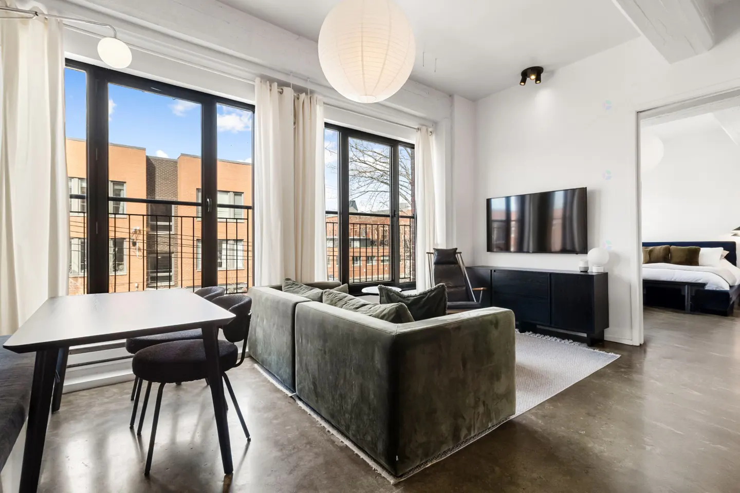 Bright living room with large windows, a gray velvet sofa, a black dining table, and a glimpse of a bedroom.