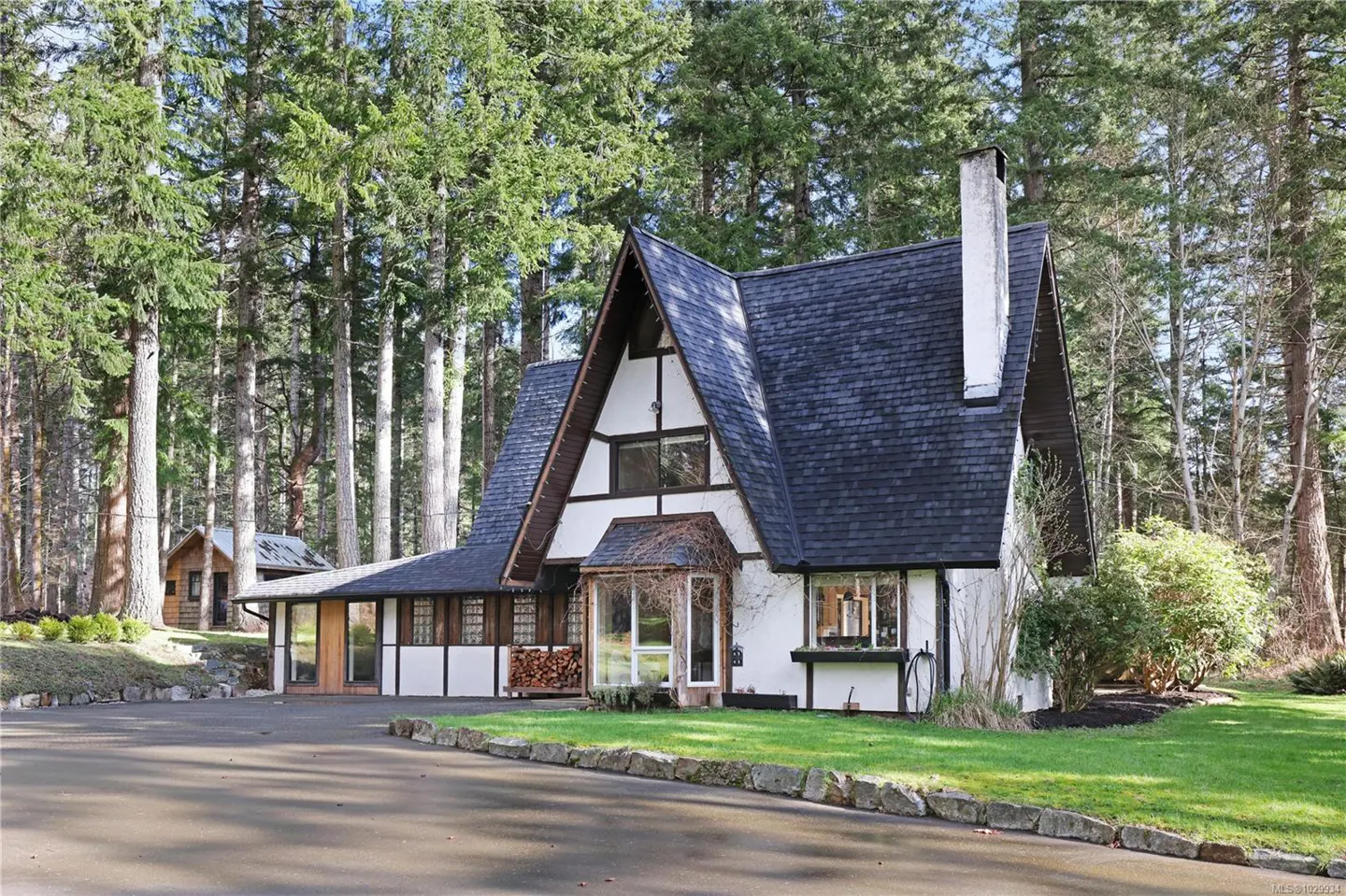 A-frame house with a dark roof, white walls, and a stone chimney, surrounded by tall trees.