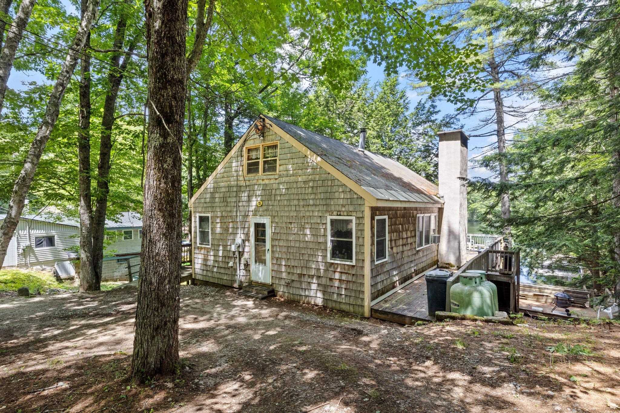 A shingled cottage with a stone chimney sits in a wooded area with a deck and propane tank visible on the side. [AI-generated text]