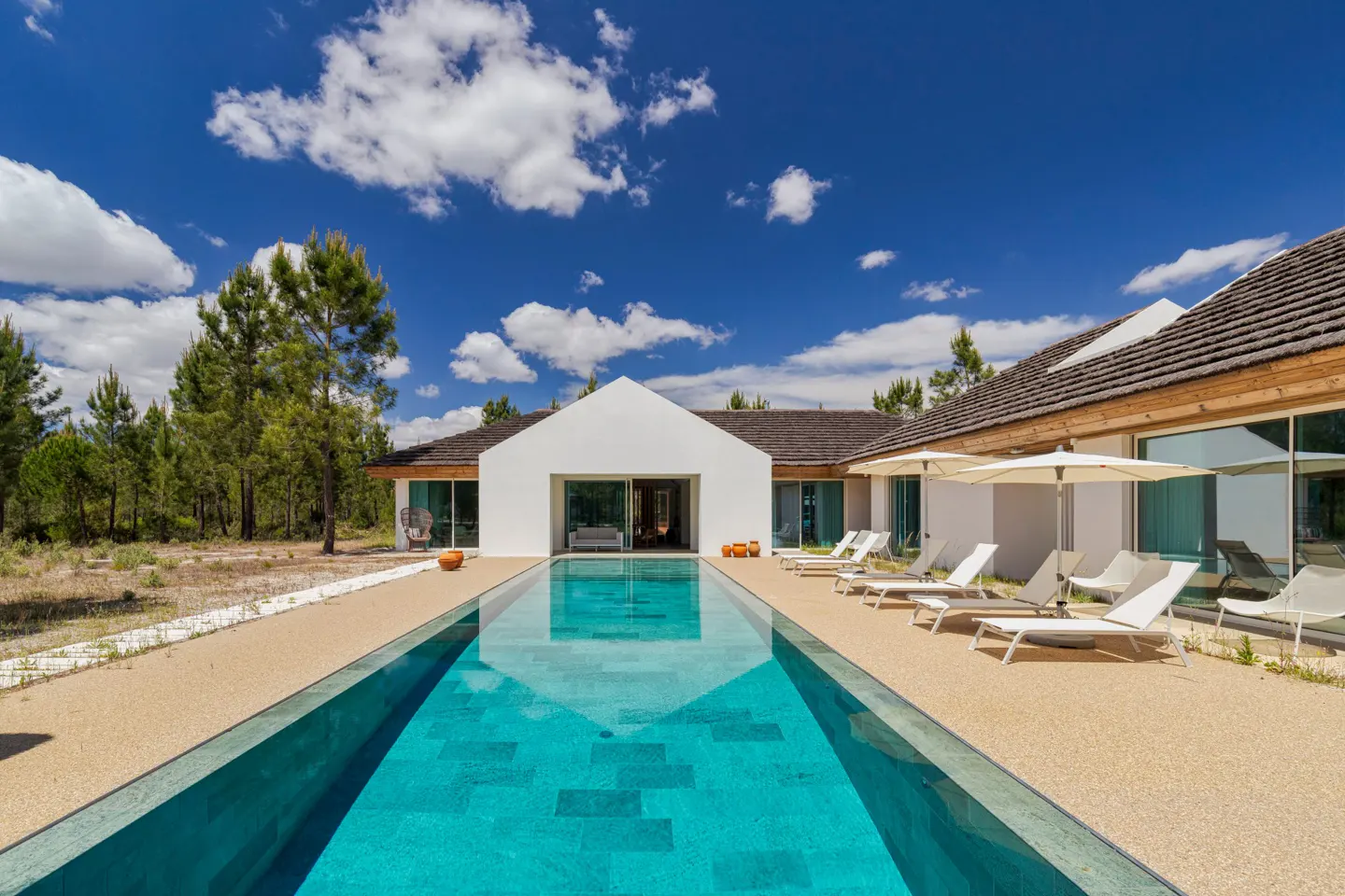 Modern white house with a long, narrow turquoise pool. Lounge chairs and umbrellas line the pool deck under a blue sky.