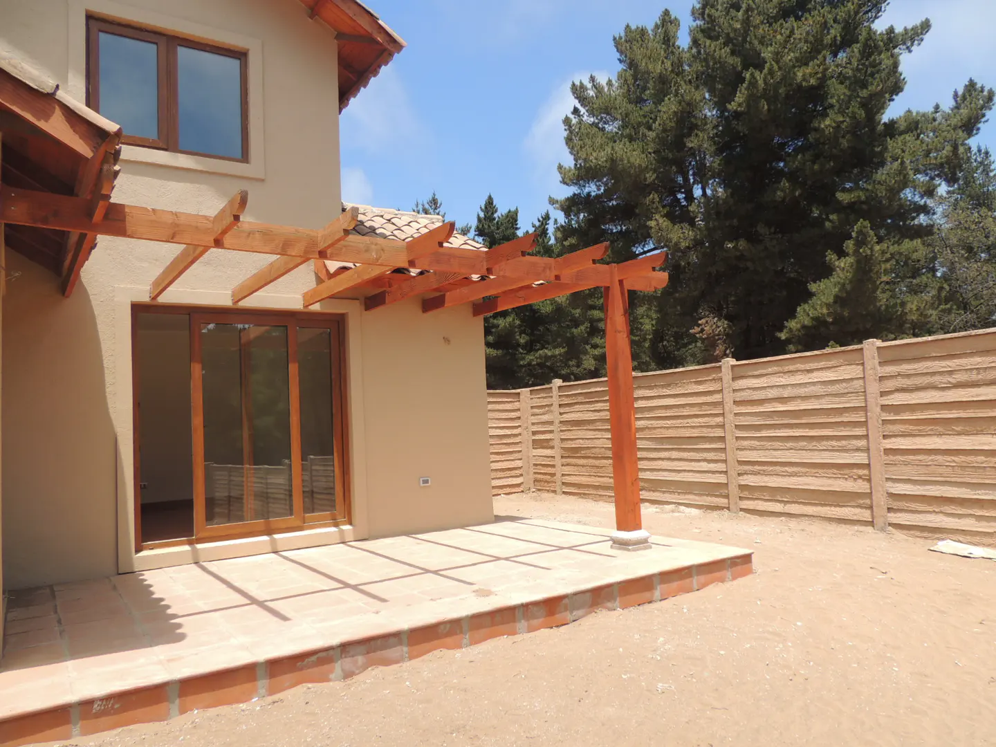 Exterior view of a beige house with a wooden pergola over a patio. A sliding glass door leads to the patio. A wooden fence and trees are in the background.
