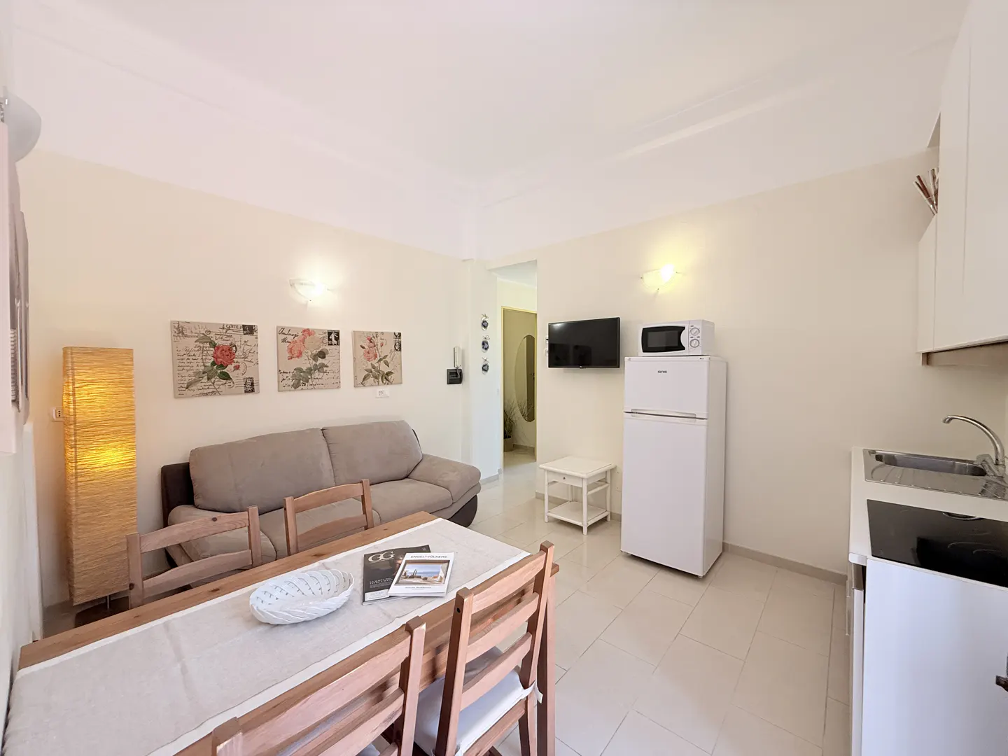 Bright studio apartment with a beige sofa, wooden dining table, and a white refrigerator and microwave. Floral art hangs on the wall.