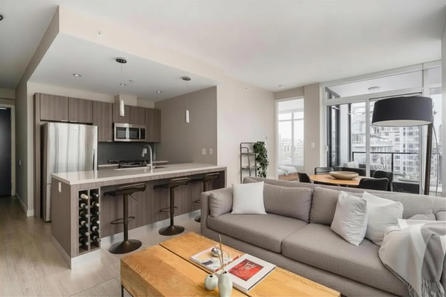 Open-concept living space with a gray sofa, wood coffee table, and kitchen island with bar stools. Large windows offer city views.