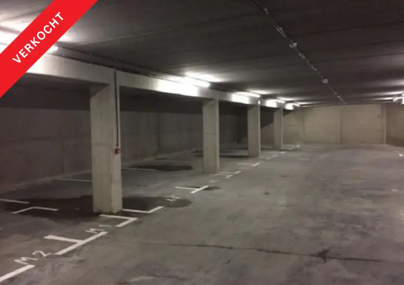 Indoor parking garage with concrete pillars and floor. White parking space lines are visible. "VERKOCHT" red banner in the upper left corner.