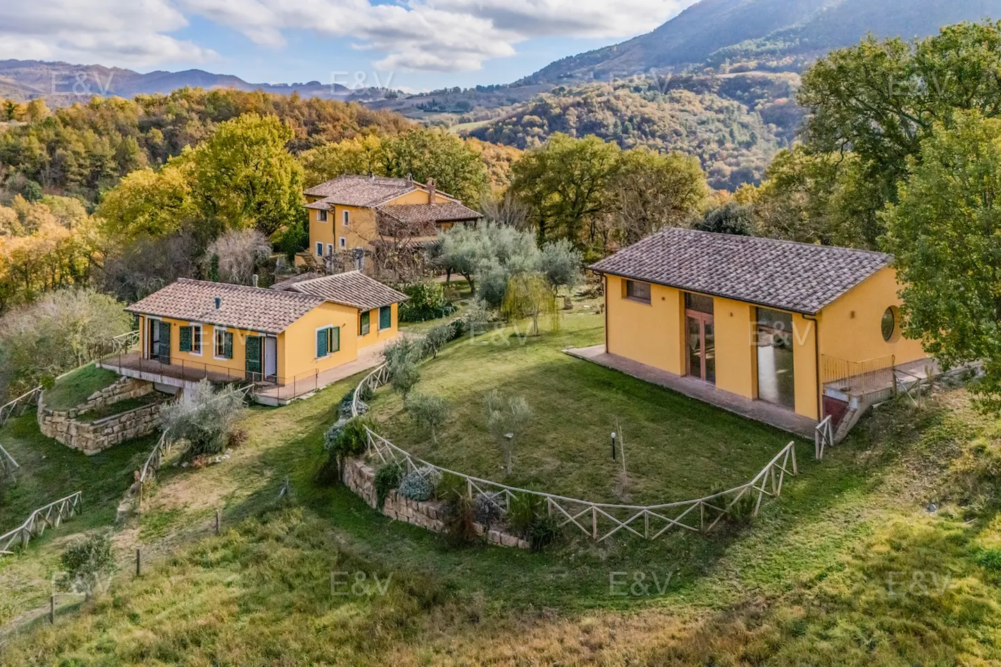 Aerial view of three yellow houses with brown roofs on a green hillside surrounded by trees and mountains.
