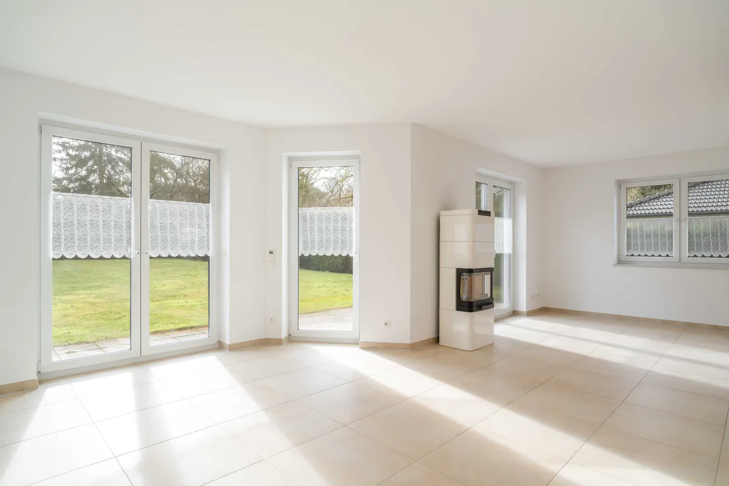 Bright, empty room with white walls, tile floor, and large windows overlooking a green lawn. A white fireplace stands in the corner.