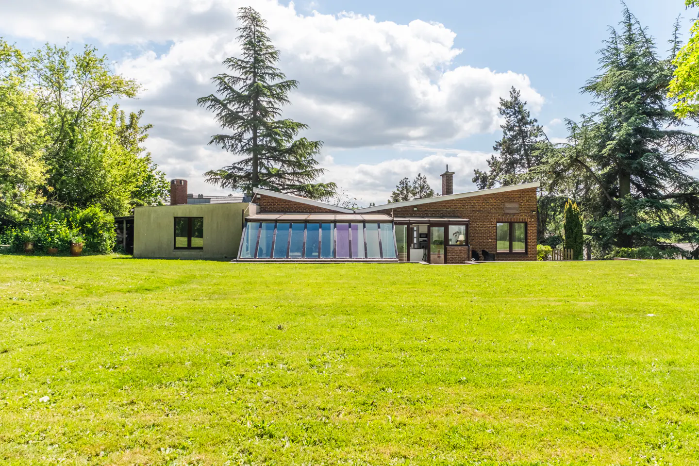 A modern brick house with a glass sunroom sits on a large green lawn under a partly cloudy sky.