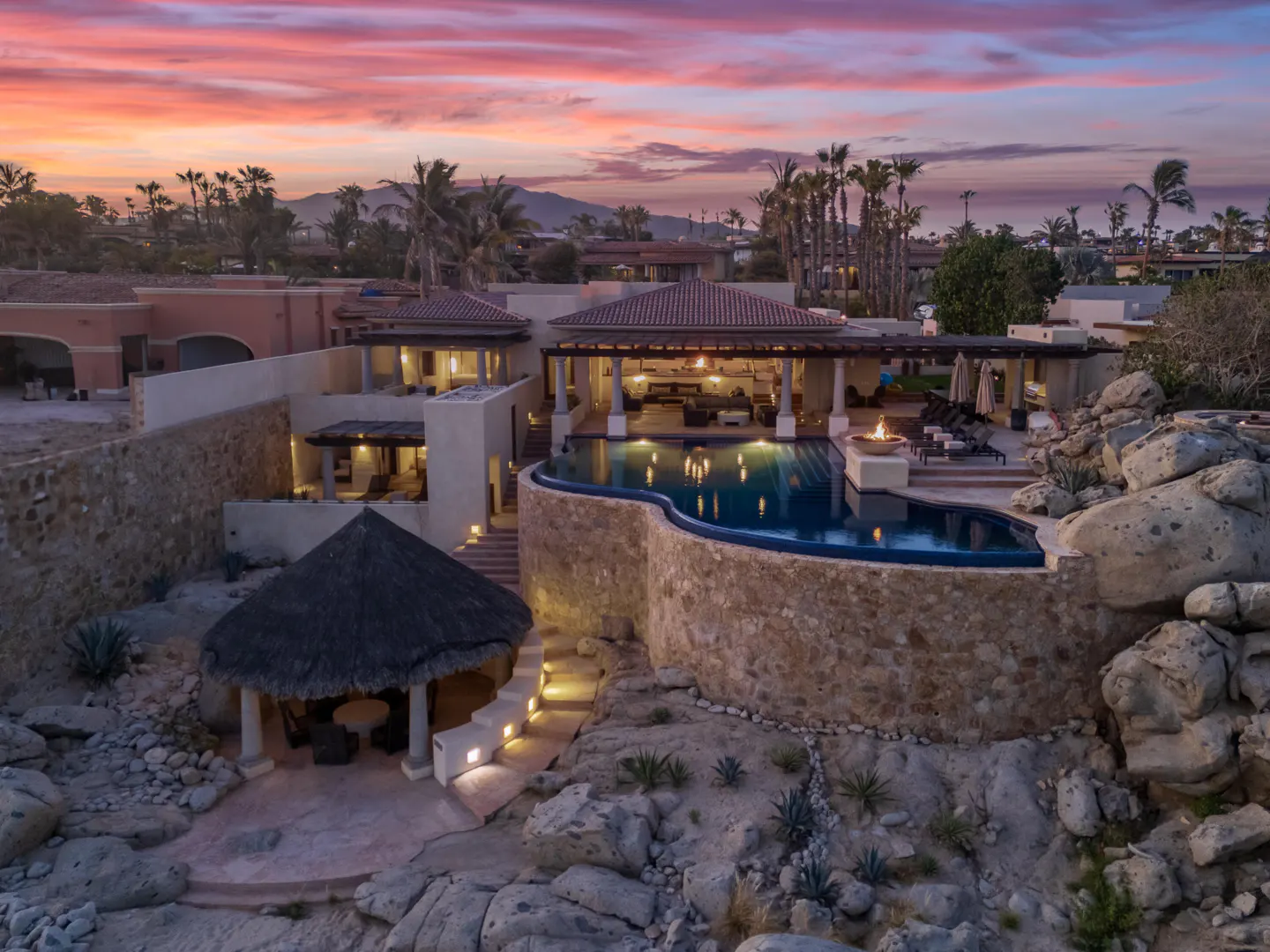 Luxury villa with infinity pool, fire pit, and thatched-roof palapa at sunset, set against a rocky desert landscape.
