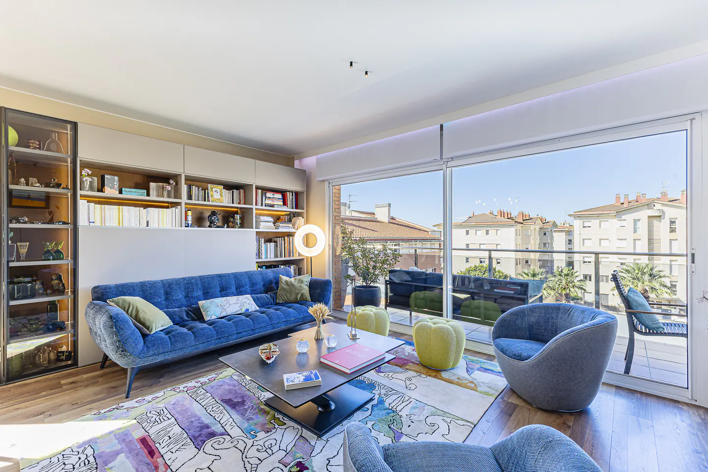 Bright living room with blue sofa, colorful rug, and floor-to-ceiling windows showing city views. Bookshelves line the wall.