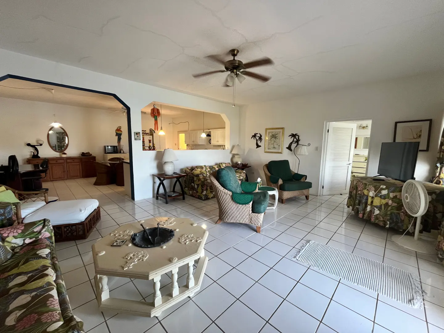 Bright living room with white tile floors, wicker furniture, floral patterns, and a ceiling fan. A kitchen is visible through an opening.