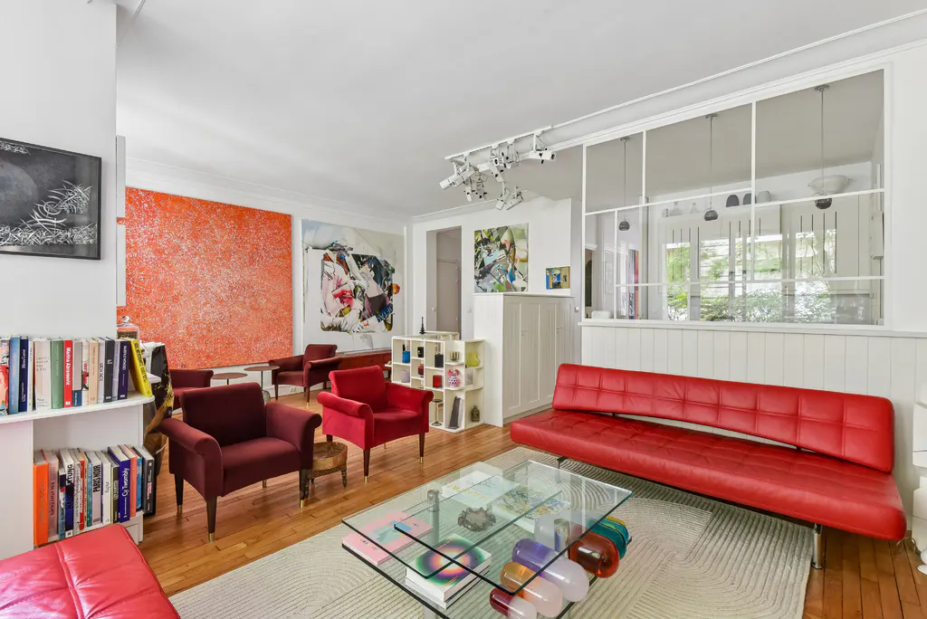 Bright living room with red sofa, armchairs, glass table, and hardwood floors. Large abstract art pieces adorn the white walls.