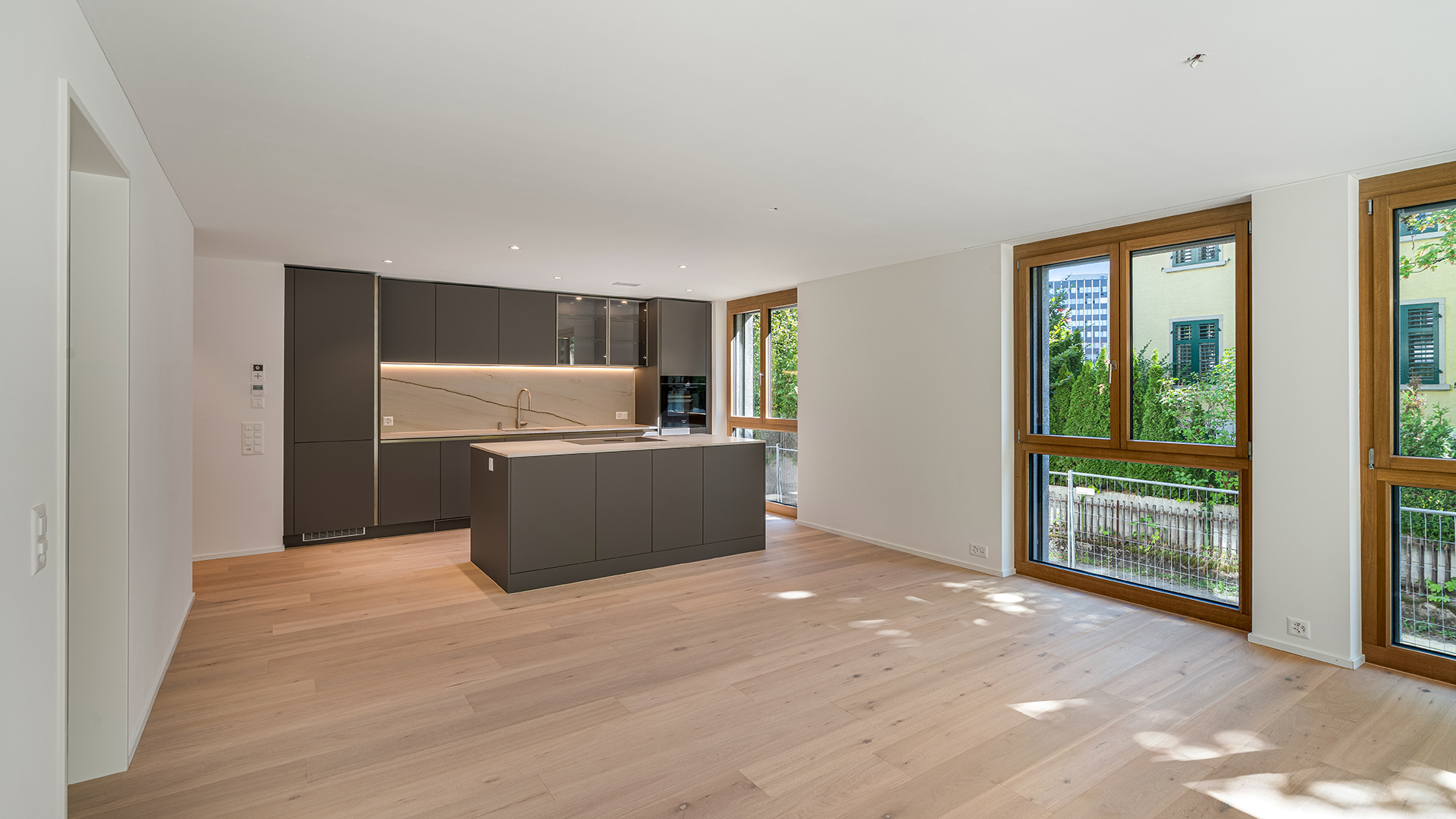 Modern kitchen with dark grey cabinets, a central island, and light wood floors, featuring large windows with wood frames. [AI-generated text]
