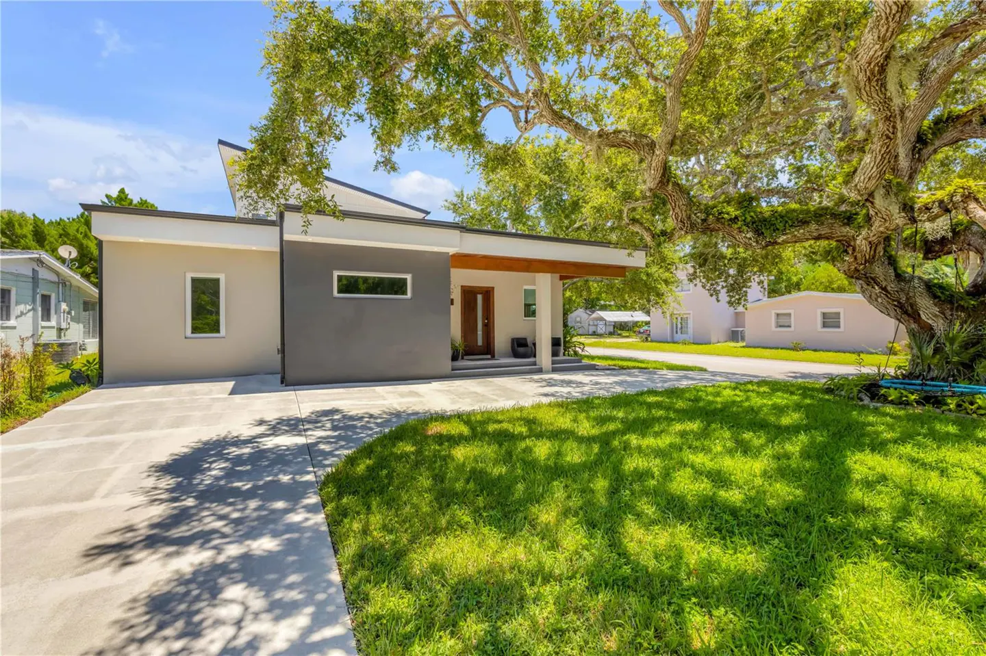Modern single-story home with gray and beige exterior, a wooden door, and a large tree in the front yard.