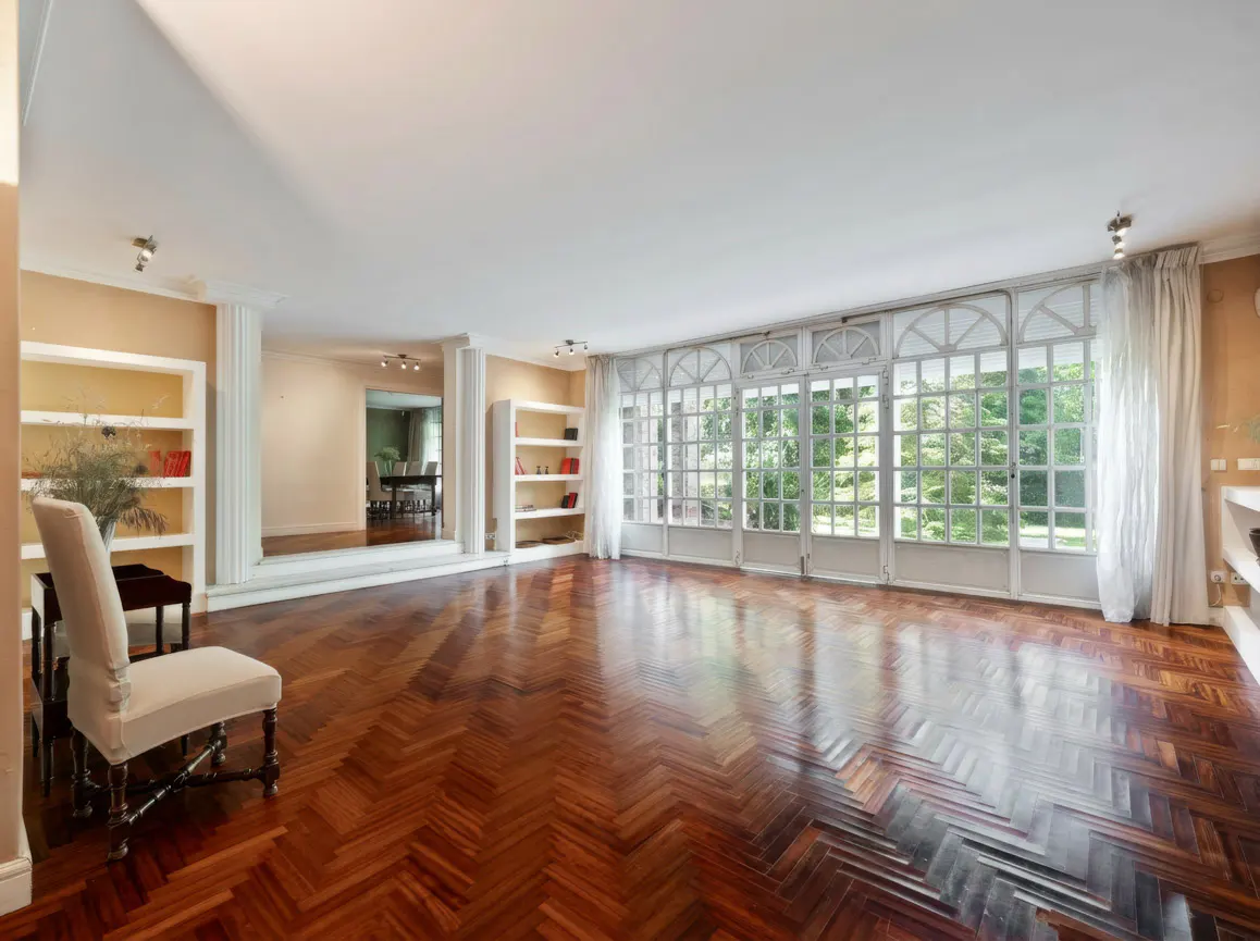 Bright, empty living room with herringbone wood floors, white walls, and large windows overlooking greenery. A chair sits near built-in bookshelves.