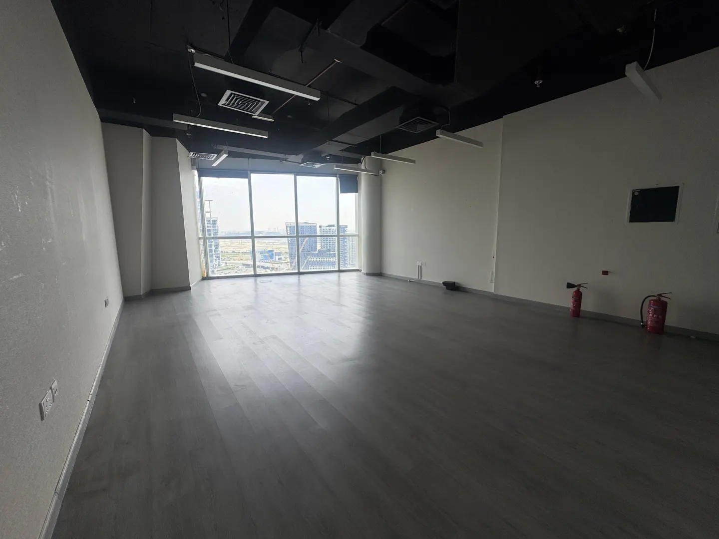 Empty office space with gray wood floors, white walls, and a black ceiling. Large windows offer a city view. Two red fire extinguishers are visible.