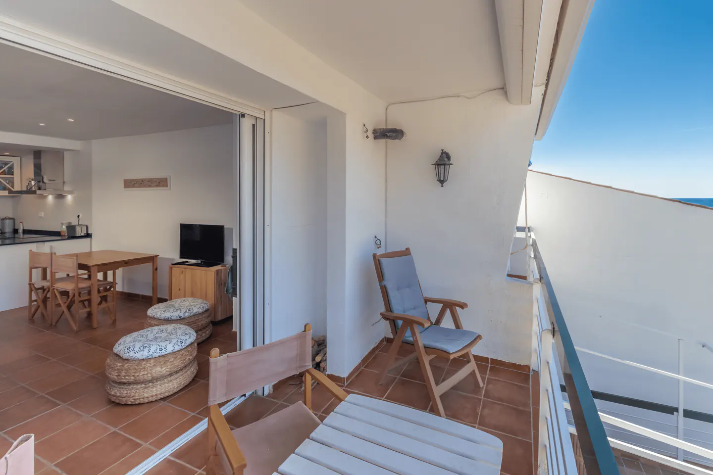 Balcony with chairs, table, and ocean view. The interior has a kitchen, dining table, and TV. The floor is tiled in brown.