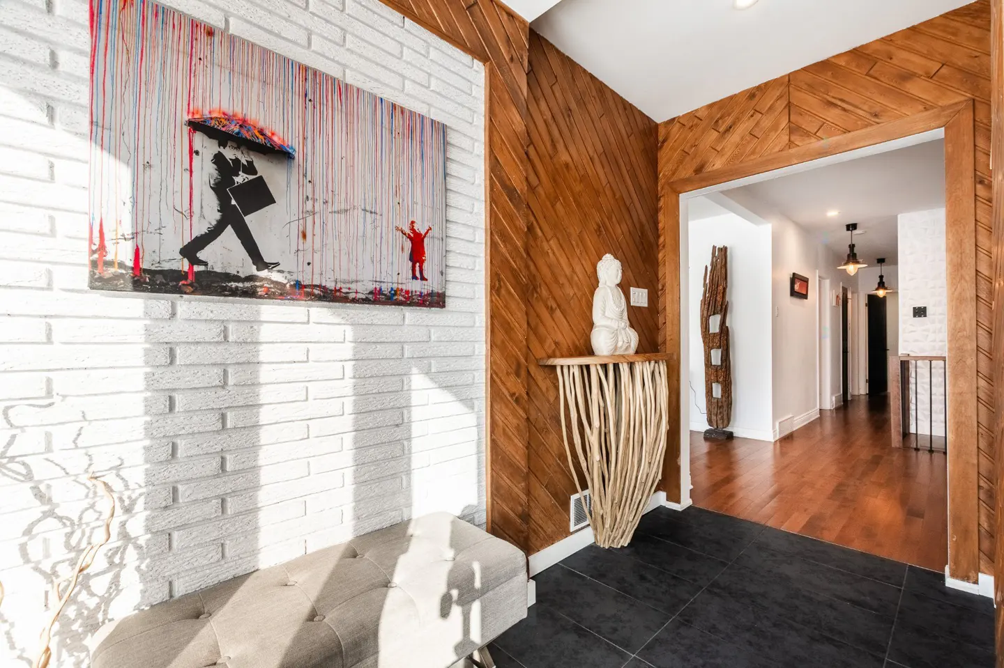 Foyer with white brick and wood paneled walls, black tile floor, and a Banksy-style art piece. A hallway is visible through an arched doorway.