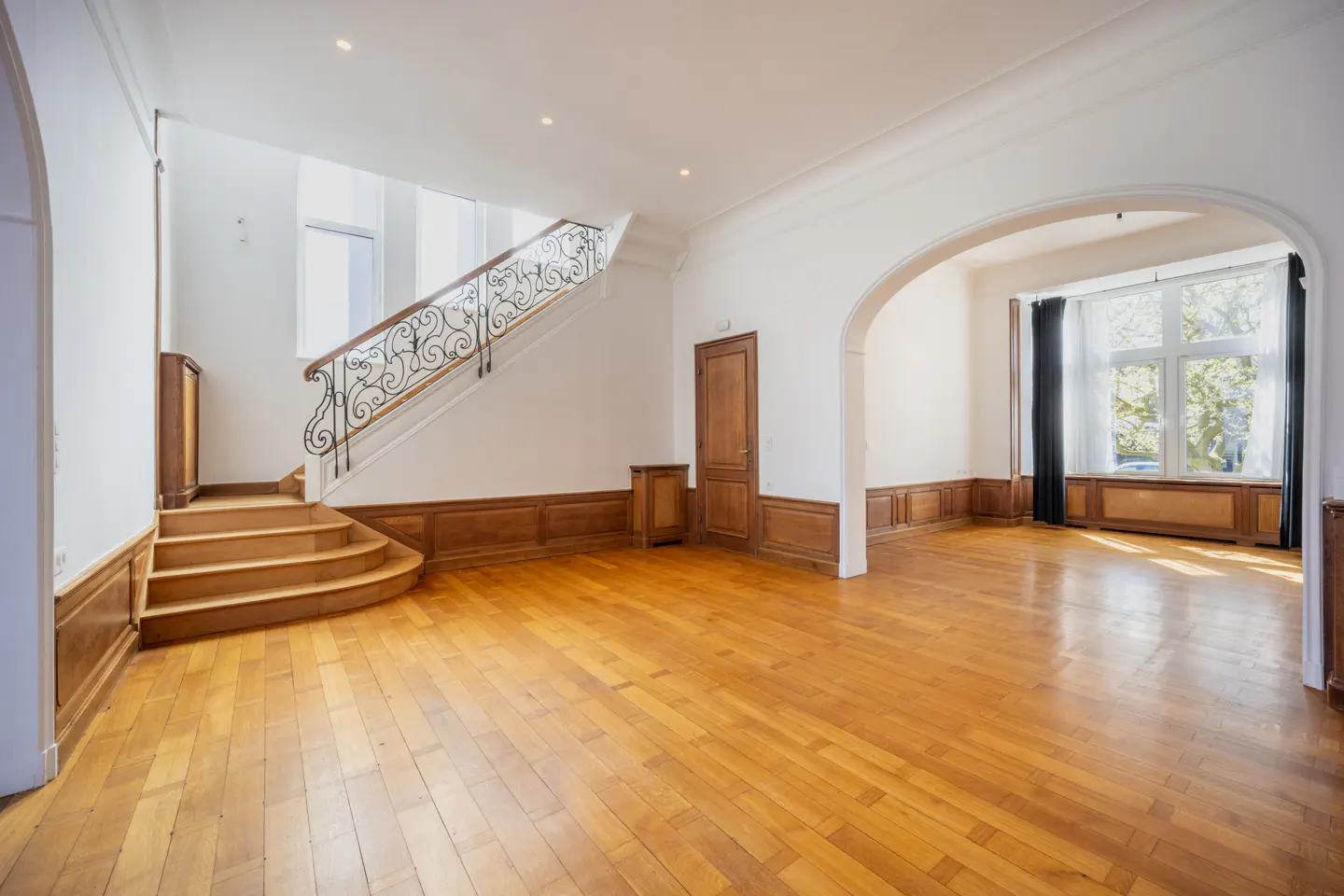 Bright, empty foyer with wood floors, white walls, and a staircase with wrought iron railing. An arched doorway leads to a window with black curtains.