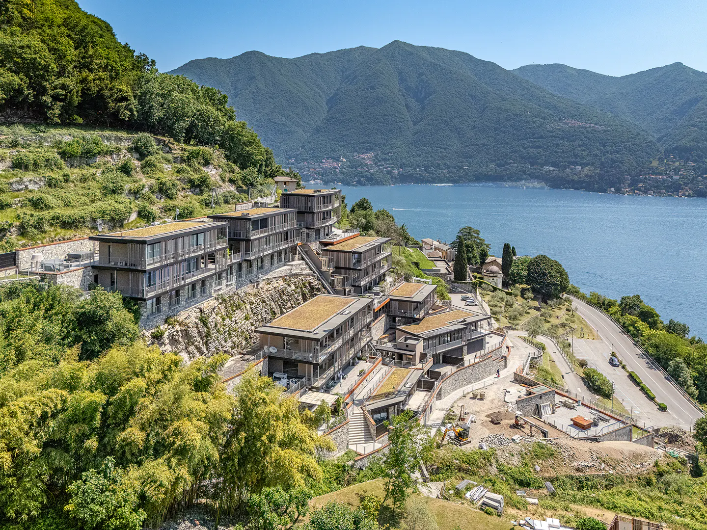 Modern terraced apartment buildings with green roofs overlooking a large lake and mountains on a sunny day.
