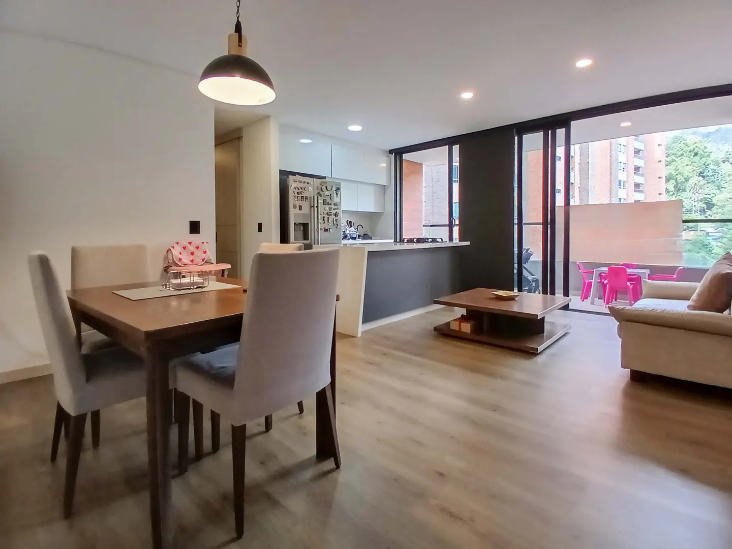 Open concept apartment with wood floors, dining table, kitchen island, and balcony with pink chairs.