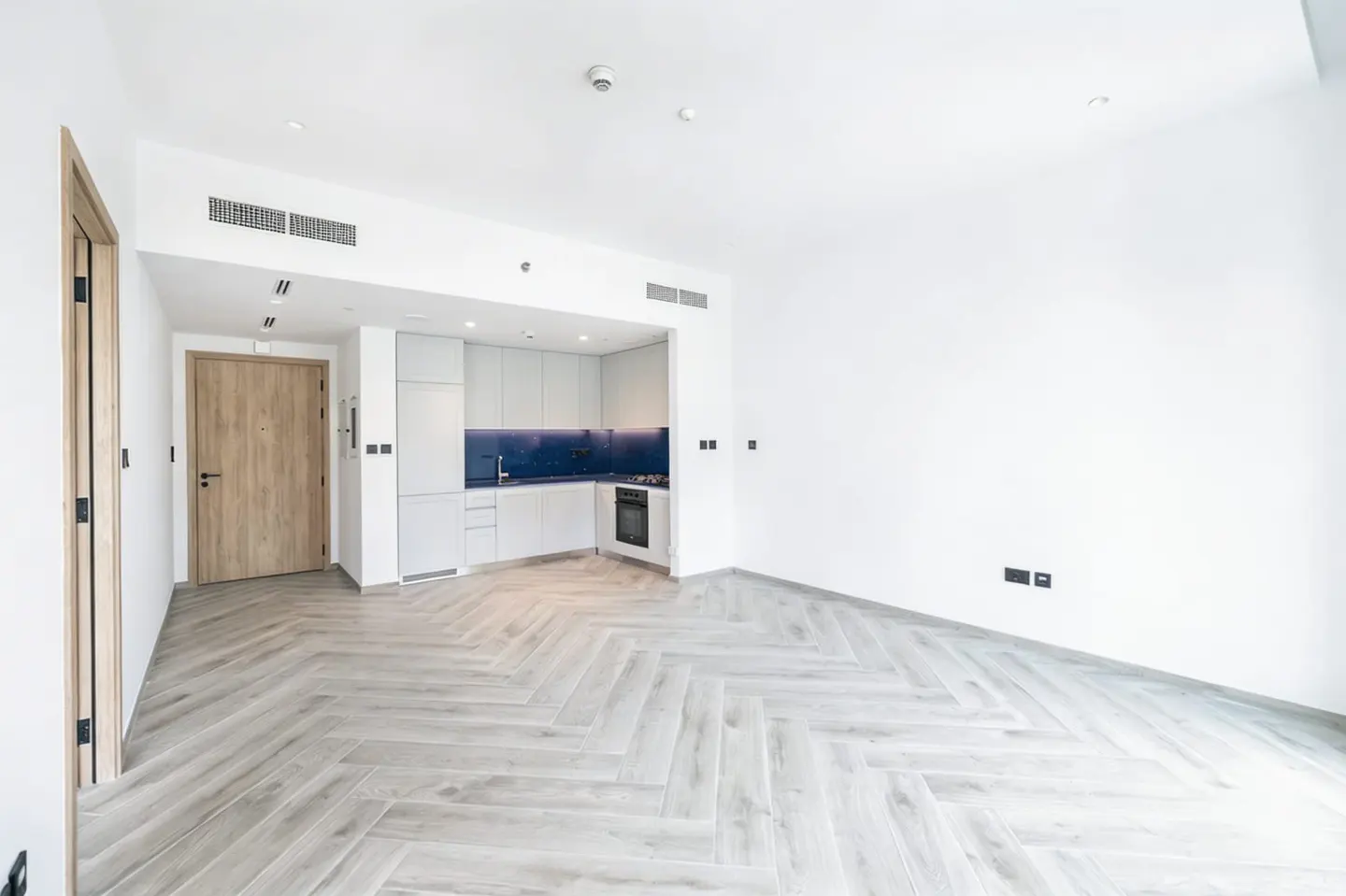 Bright, empty apartment with white walls, herringbone wood floors, and a modern white kitchen with blue backsplash.