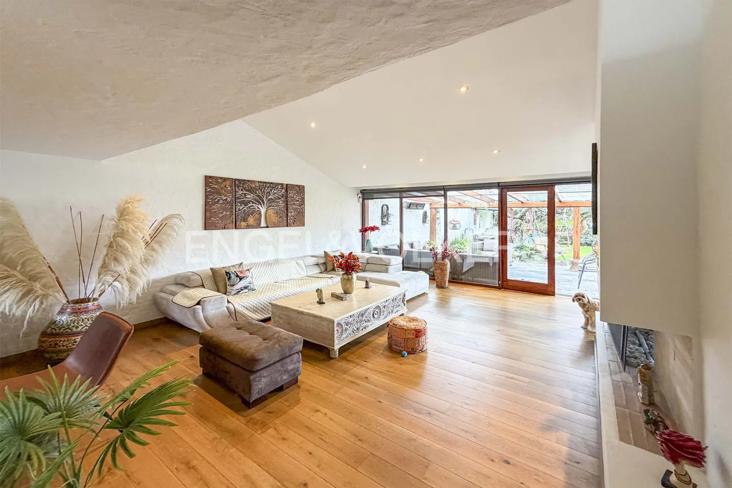 Bright living room with hardwood floors, white walls, and a large sectional sofa. Sliding glass doors lead to an outdoor patio. A small dog stands near the fireplace.