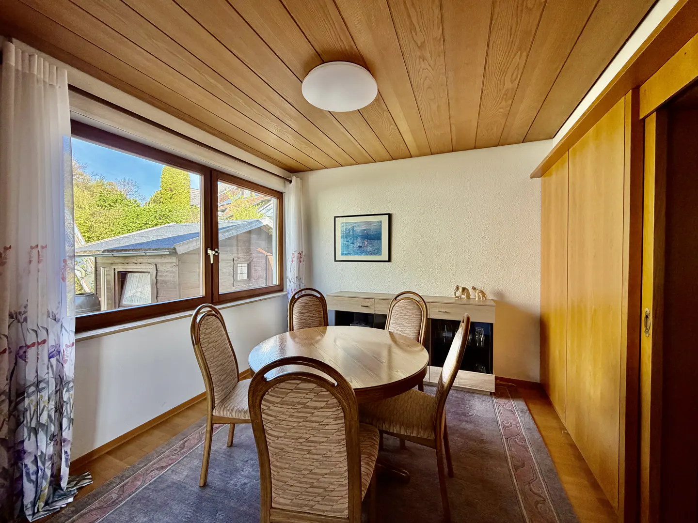 A dining room with a round wooden table and six chairs, a wooden ceiling, and a window with a view of a garden.