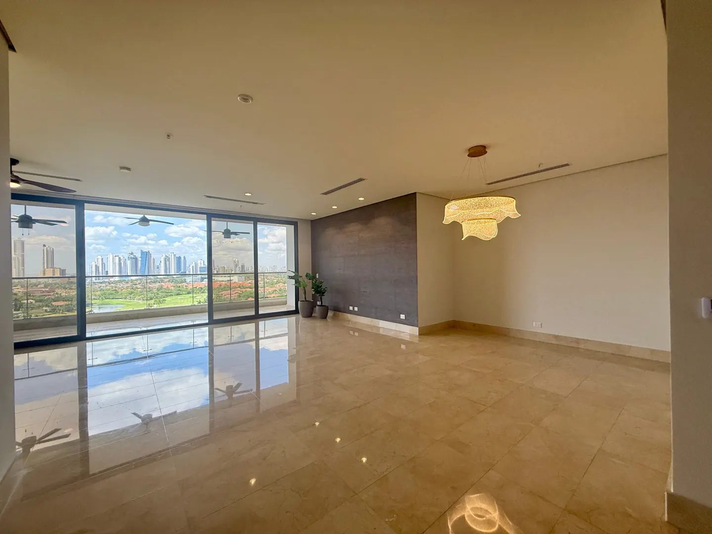 A spacious, empty room with marble floors and a large window overlooking a city skyline. A modern chandelier hangs from the ceiling.