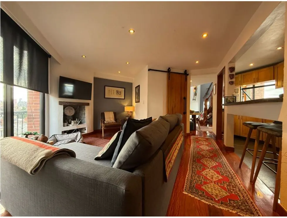 Living room with gray sofa, fireplace, TV, and a red patterned rug on a hardwood floor. Kitchen with bar stools is visible.