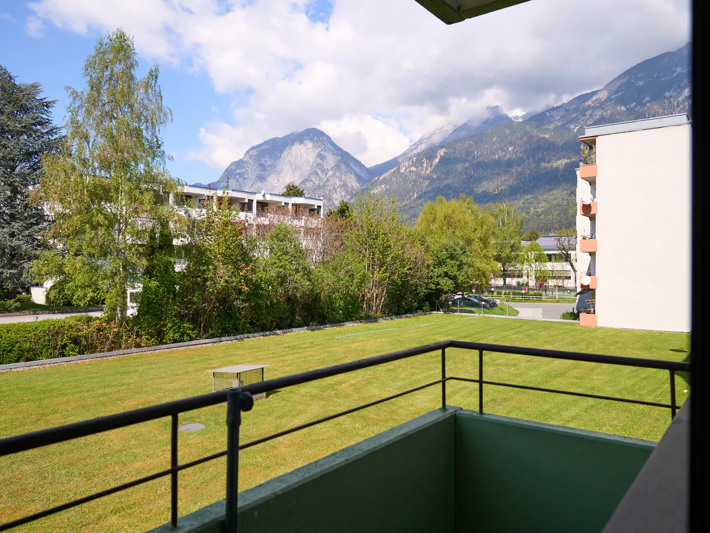 View from a balcony with black railings overlooking a green lawn, trees, buildings, and mountains under a cloudy sky.