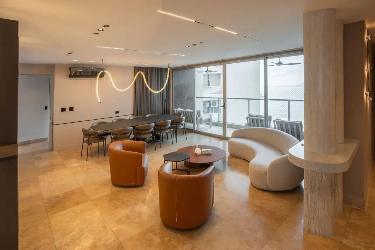 Open-concept living and dining area with tan tile floors, brown leather chairs, and a long black dining table. A curved white sofa faces a balcony.