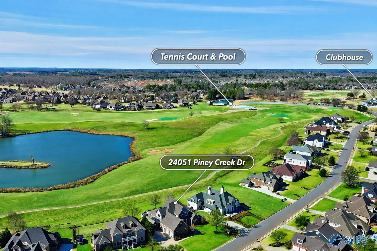 Aerial view of a residential area with houses, a golf course, a lake, a tennis court, a pool, and a clubhouse under a blue sky.
