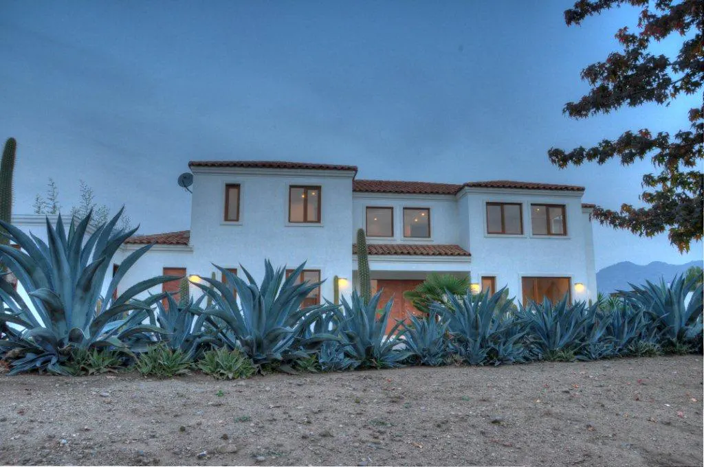 Two-story white house with a red tile roof, surrounded by blue agave plants and cacti.