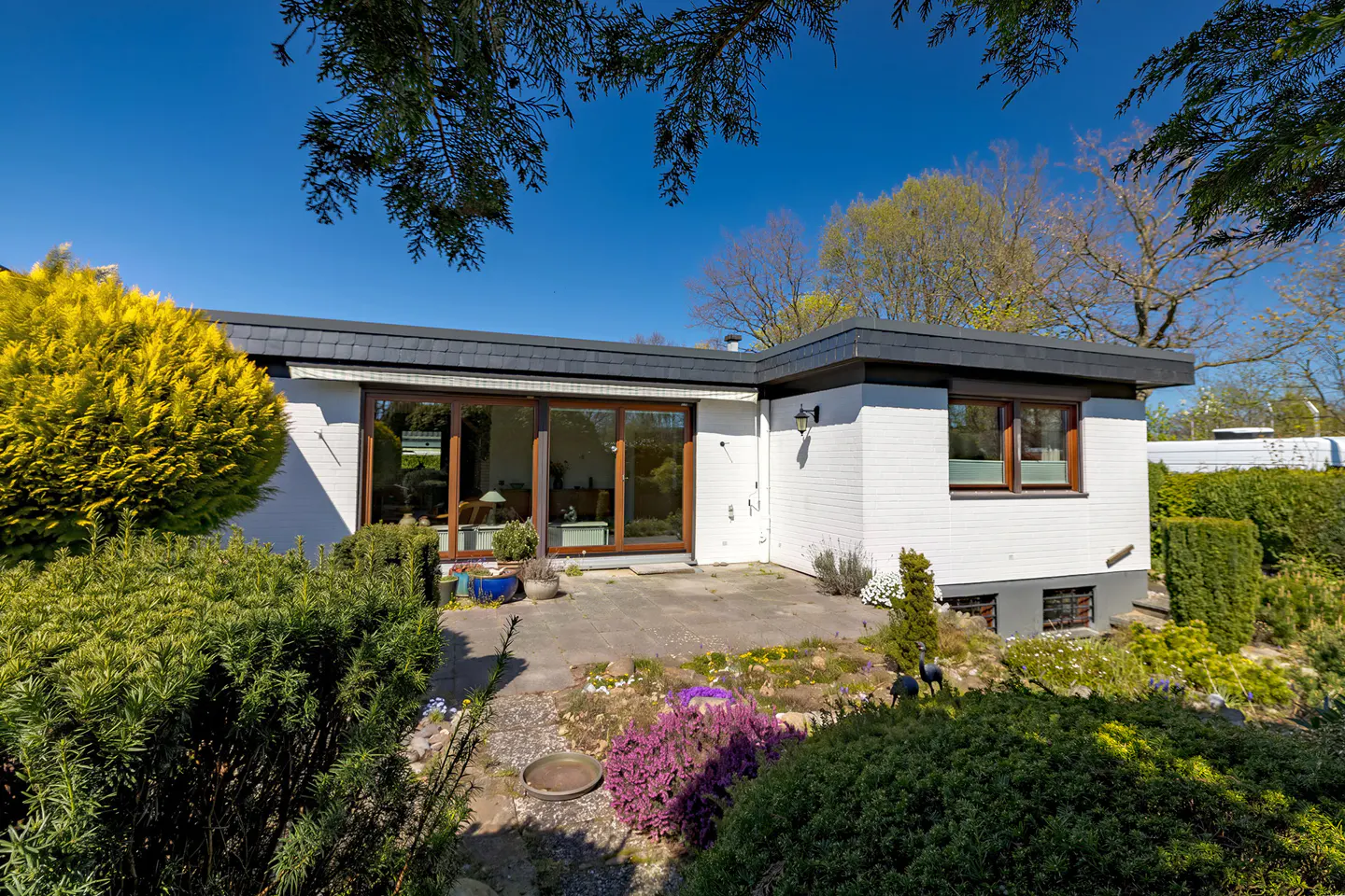 Exterior view of a white brick house with a dark gray roof, surrounded by lush green landscaping and a blue sky.
