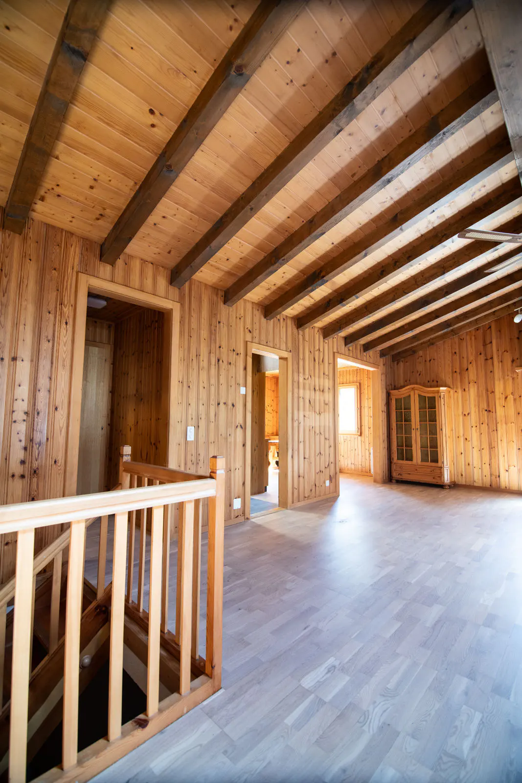 Interior view of a wood-paneled room with exposed beams, light wood floors, and a staircase with a wooden railing.