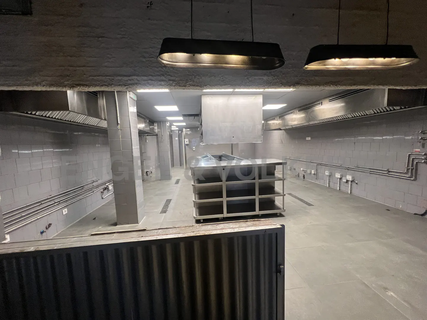 A commercial kitchen with stainless steel appliances, gray tiled walls, and overhead lighting.