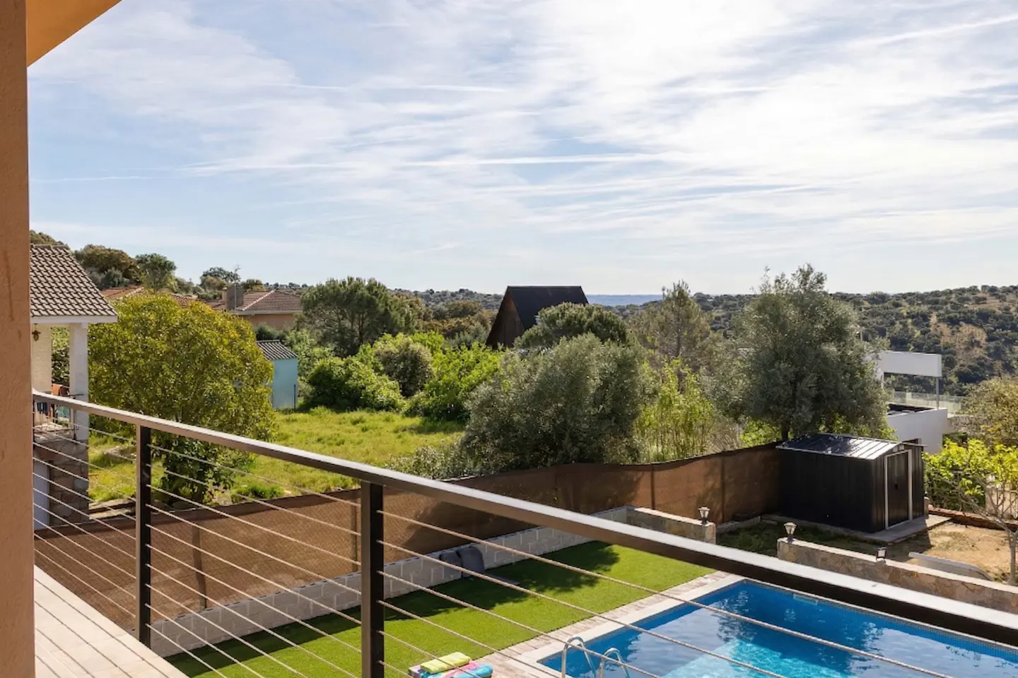 View from balcony with metal railing overlooking a blue swimming pool, green lawn, trees, and houses under a blue sky.