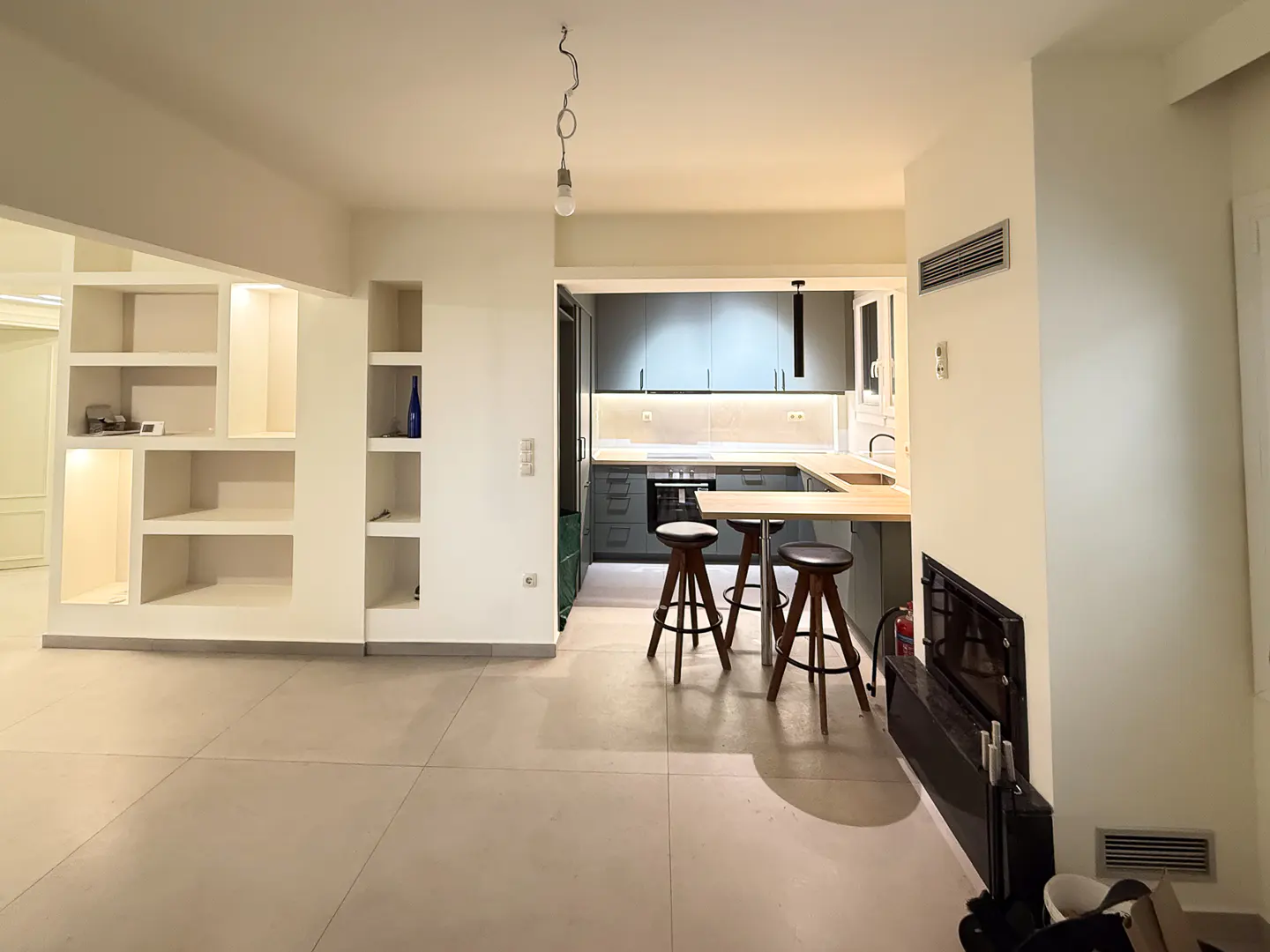 Open-concept living space with a gray kitchen, wood breakfast bar, and three stools. White built-in shelves and a black fireplace are visible.